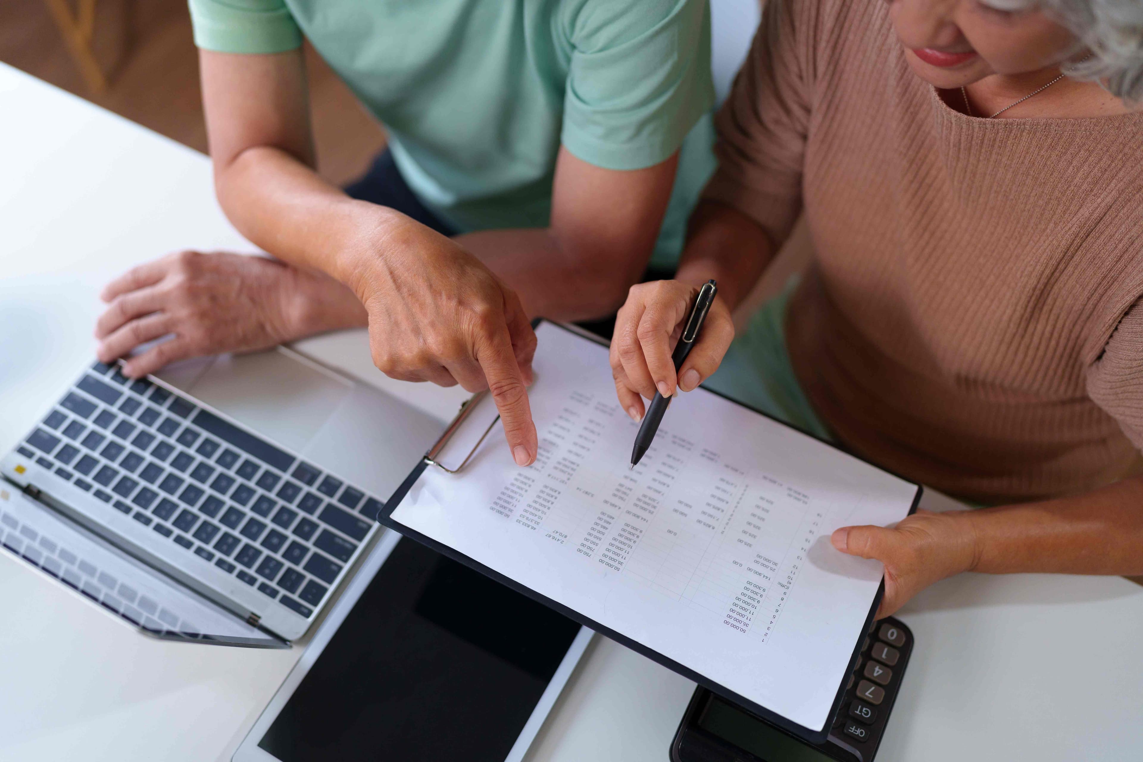 Two people at a desk reviewing a financial spreadsheet on a clipboard with a calculator, laptop, and tablet nearby.